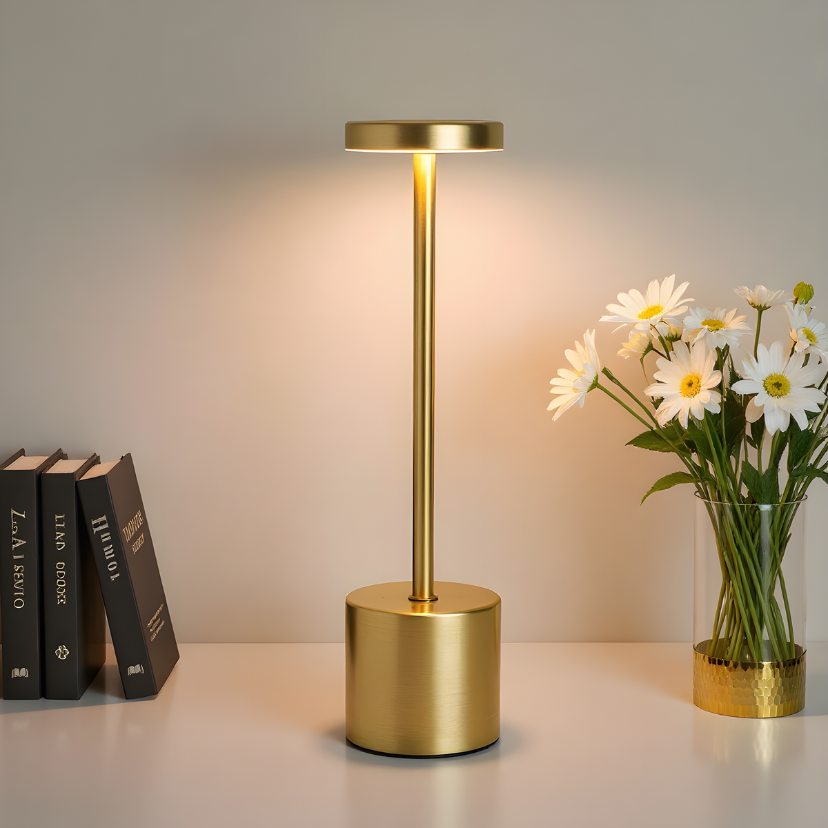 Lampe de table LED dorée allumée, posée entre des livres et un vase de fleurs blanches.